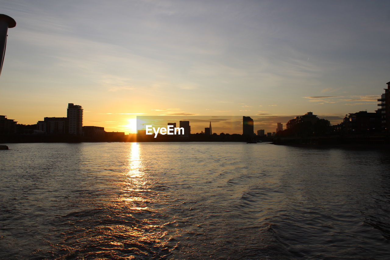 SILHOUETTE BUILDINGS BY RIVER AGAINST SKY AT SUNSET