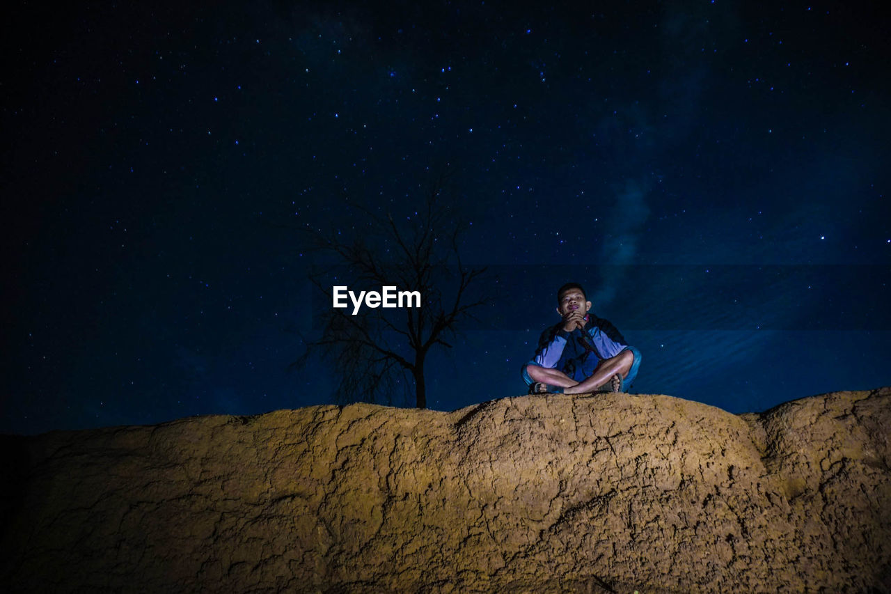Low angle view of man sitting on rock formation against star field