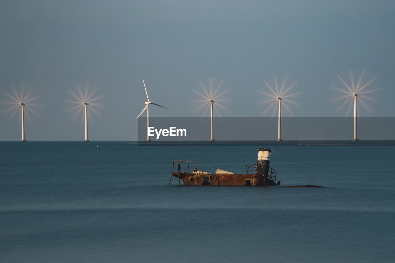 Abandoned boat by wind turbines on sea shore against sky