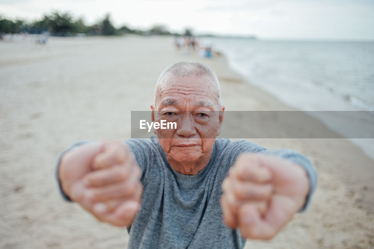 Portrait of senior man exercising while standing at beach against sky during sunset