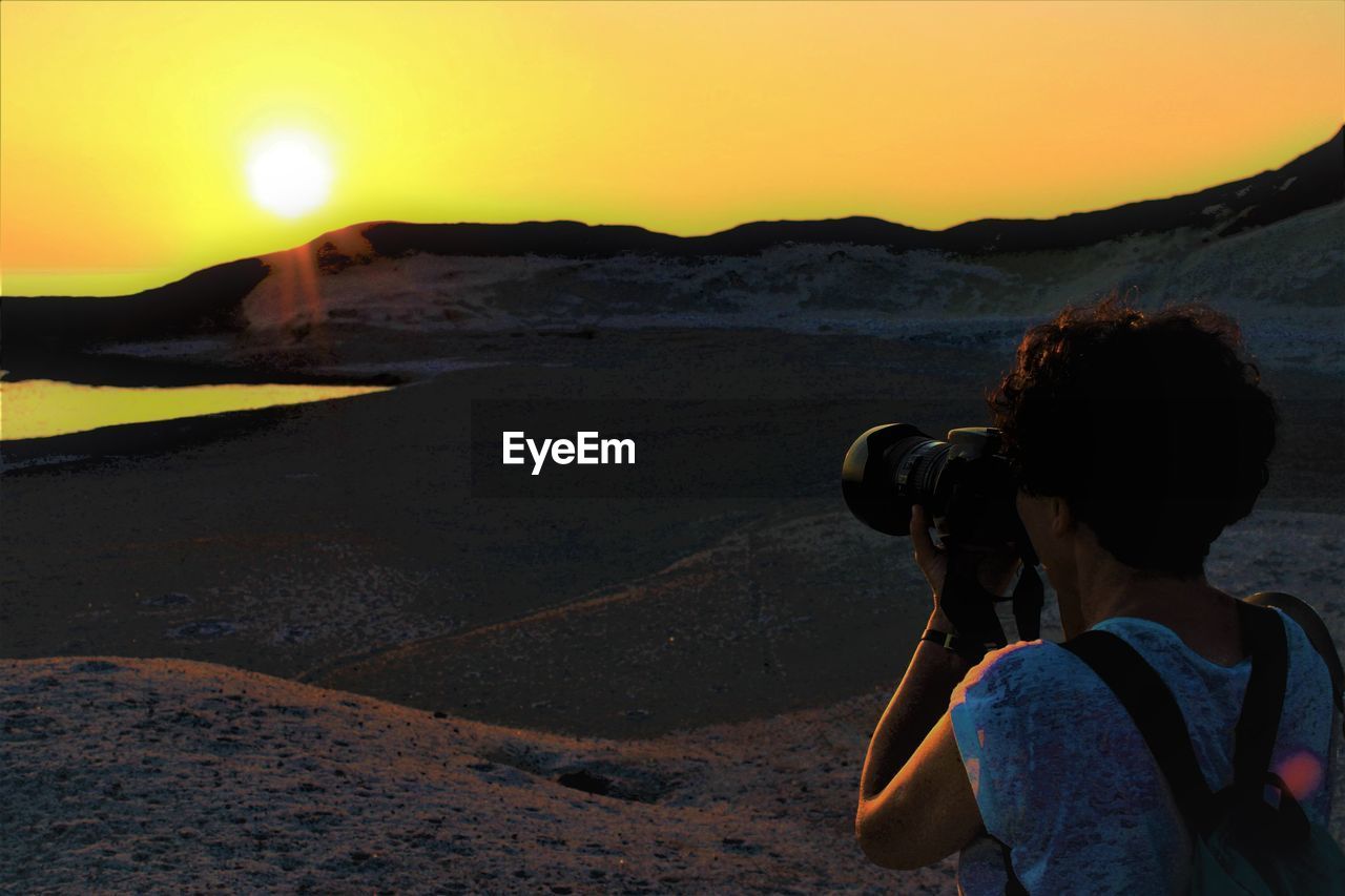 Woman photographing at beach against sky during sunset