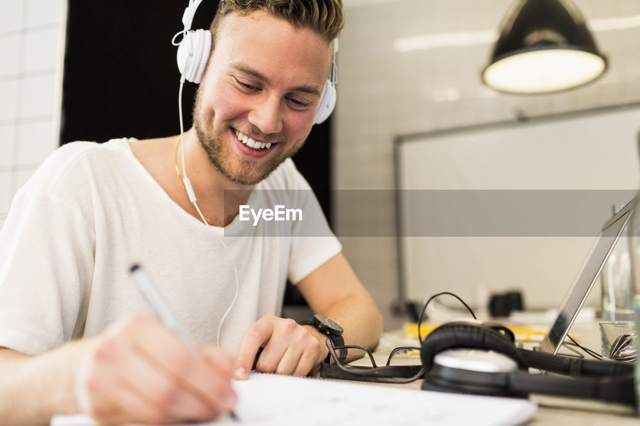 Happy young businessman wearing headphones while writing in book at creative office