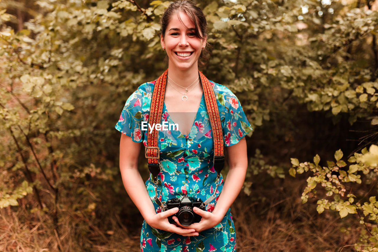 Portrait of smiling young woman with camera standing against tree