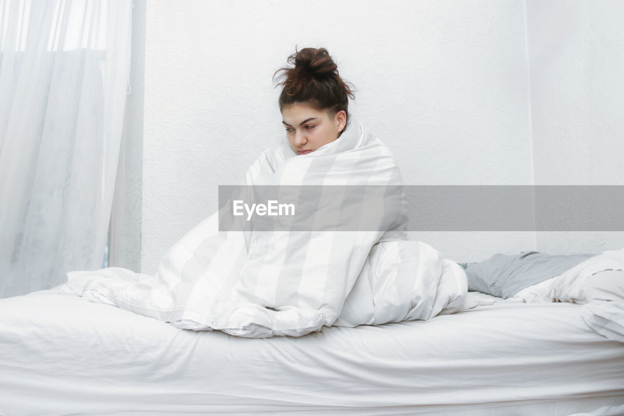 bed, white, one person, bedroom, indoors, furniture, women, domestic room, adult, relaxation, linen, lifestyles, young adult, dress, sitting, female, sheet, person, gown, full length, textile, morning, pillow, clothing, looking, bathrobe, front view, bride