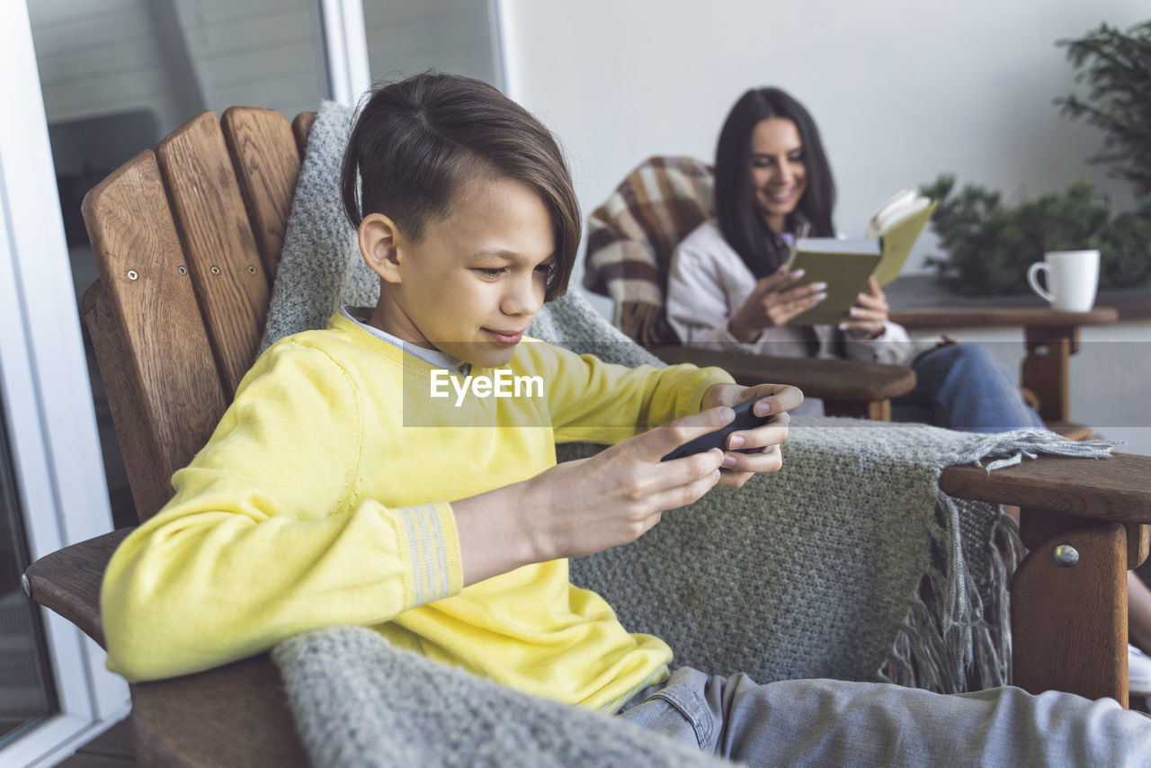 Boy using smart phone while mother reading book on balcony