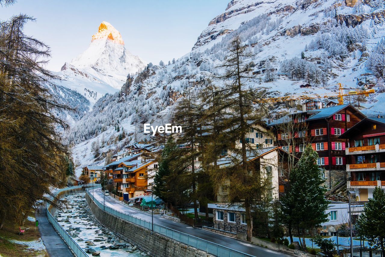 SNOW COVERED ROAD BY BUILDINGS AGAINST SKY