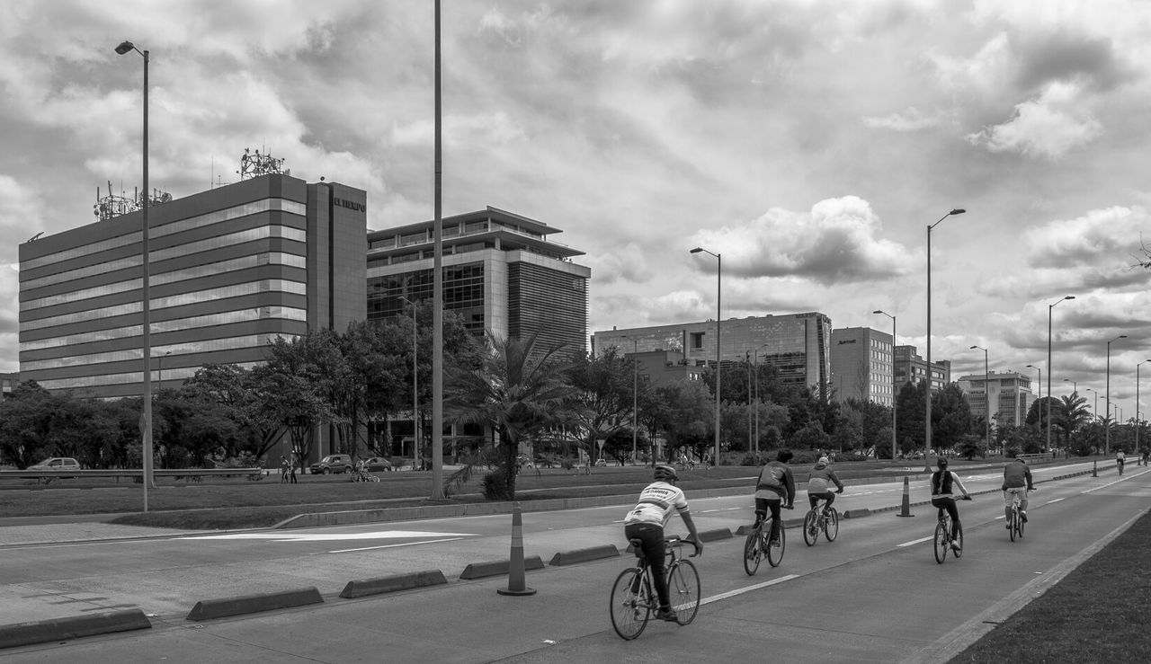 Men cycling on road by buildings against cloudy sky