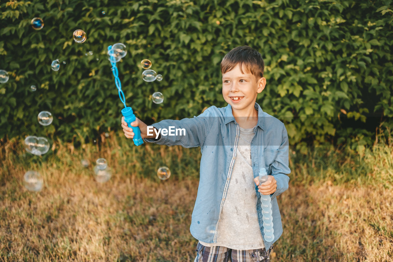 A boy is playing with soap bubbles in a summer park, among the greenery.