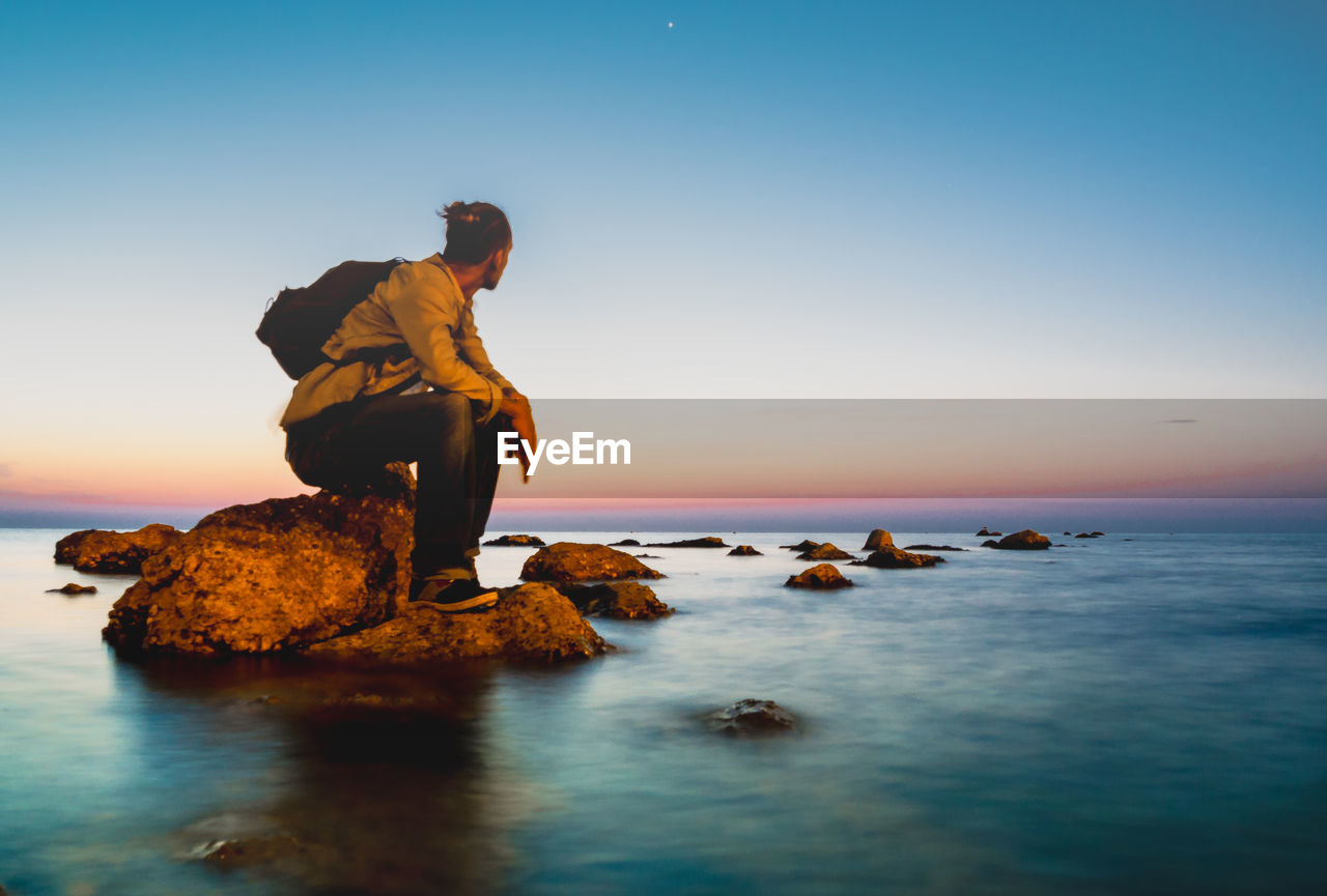 Man sitting on rock by sea against sky during sunset