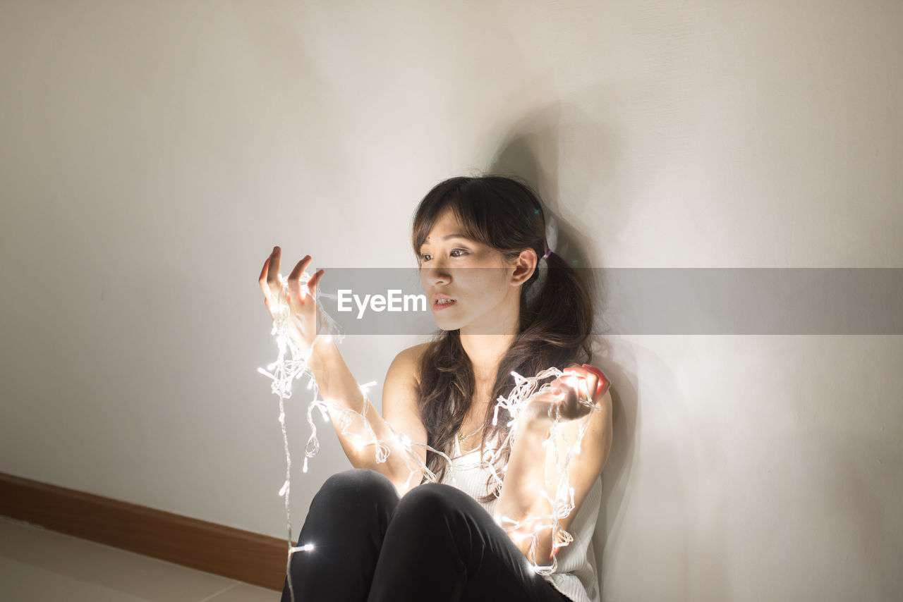 Young woman with illuminated string light sitting by wall at home