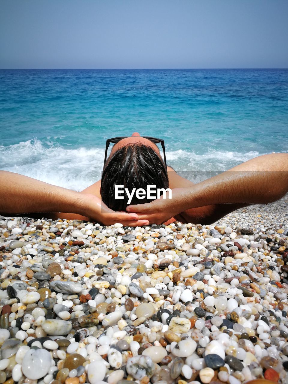 Man lying on pebbles shore at beach against sky during sunny day