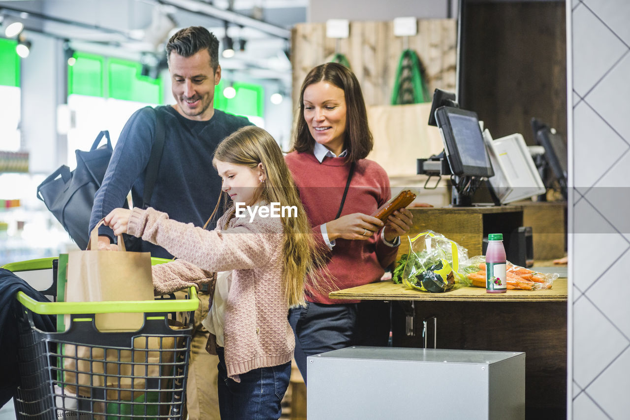 Smiling parents looking at daughter arranging paper bags in shopping cart