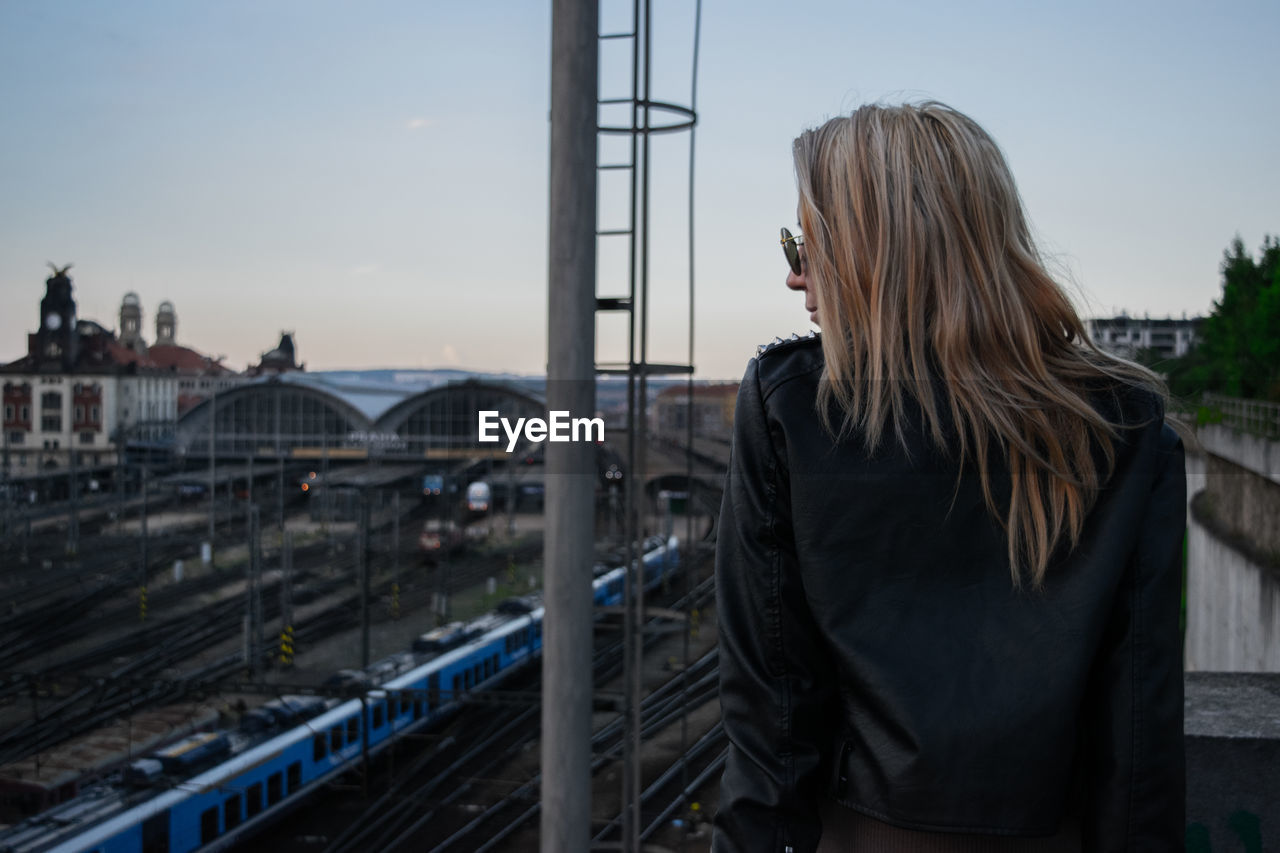 Young woman with blond hair standing by shunting yard against sky during sunset