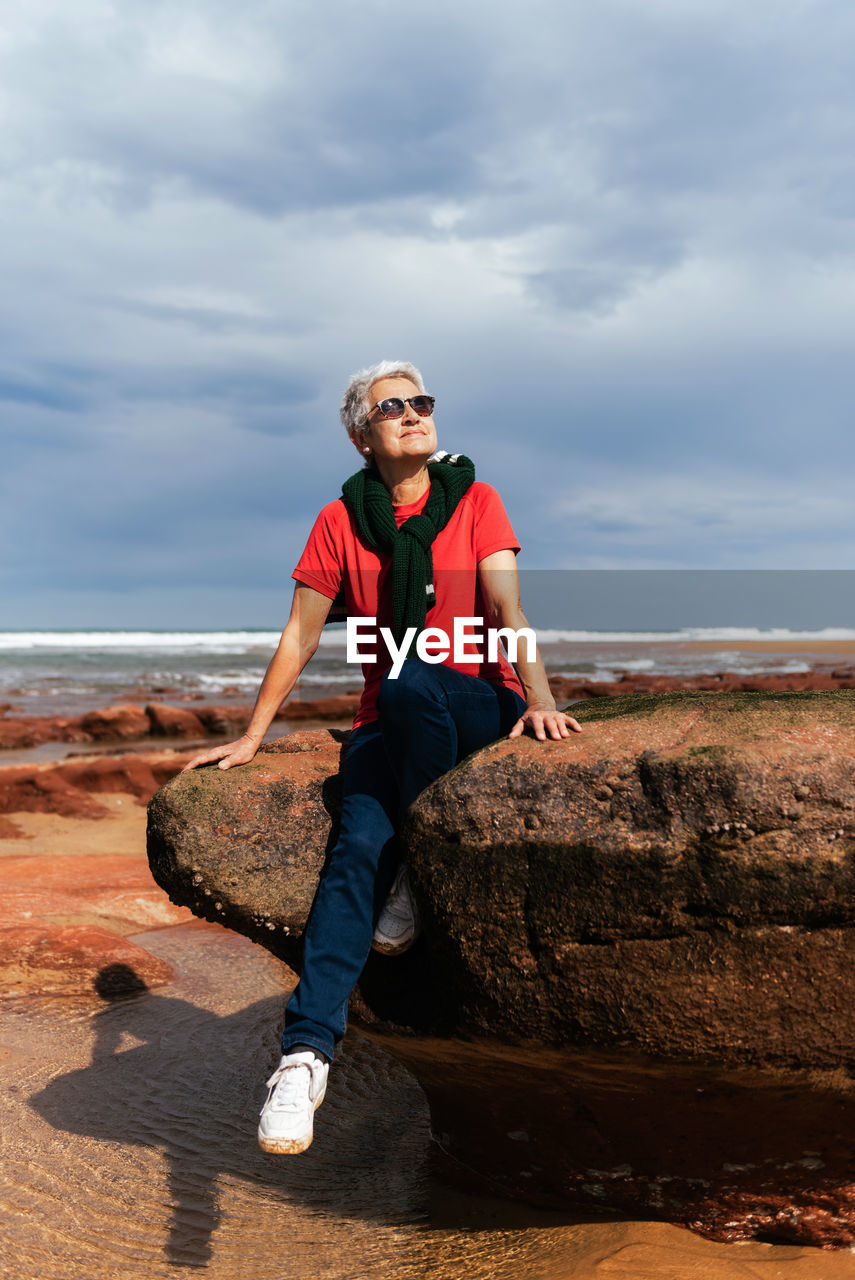 Smiling elderly female tourist in sunglasses sitting on rough boulder while looking up against ocean under cloudy sky