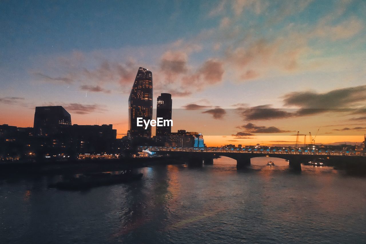 Illuminated bridge over river by buildings against sky during sunset