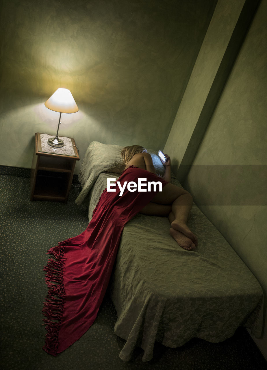 Naked woman wrapped in blanket using phone while lying on bed at home