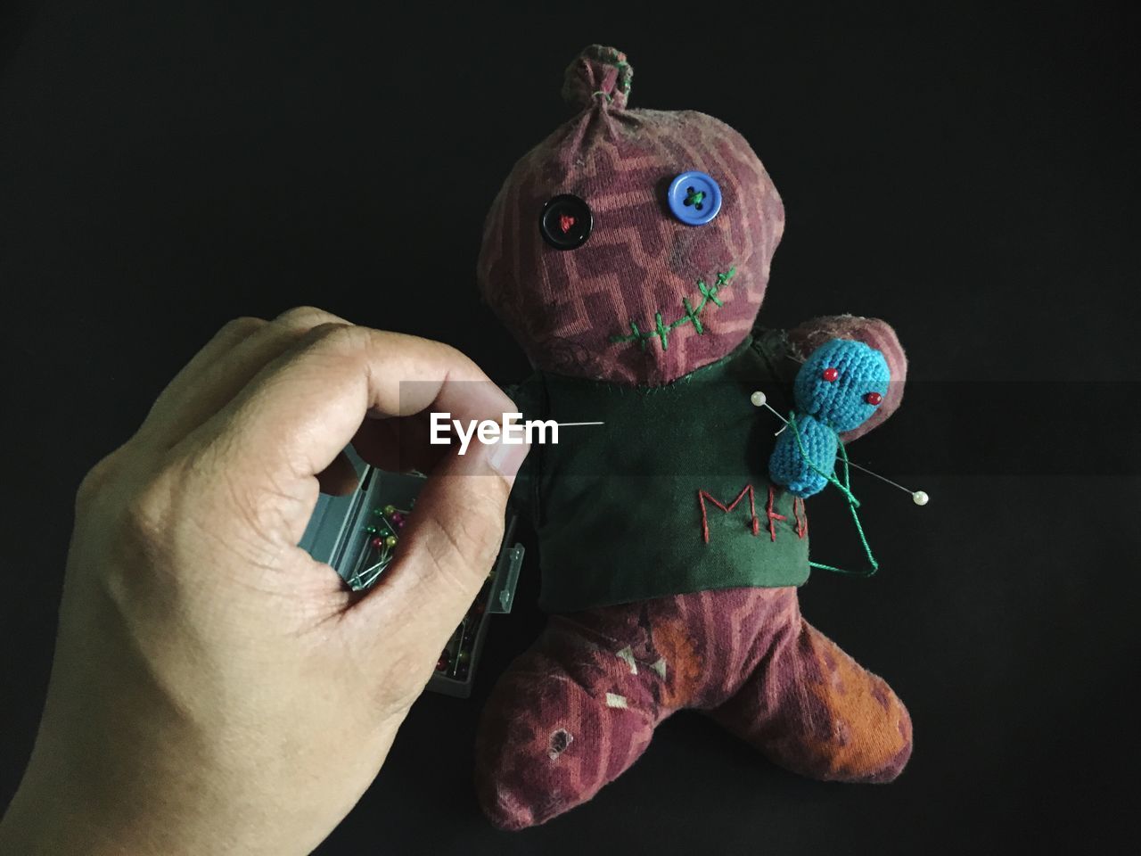 Cropped hand of man putting straight pin in voodoo doll over black background