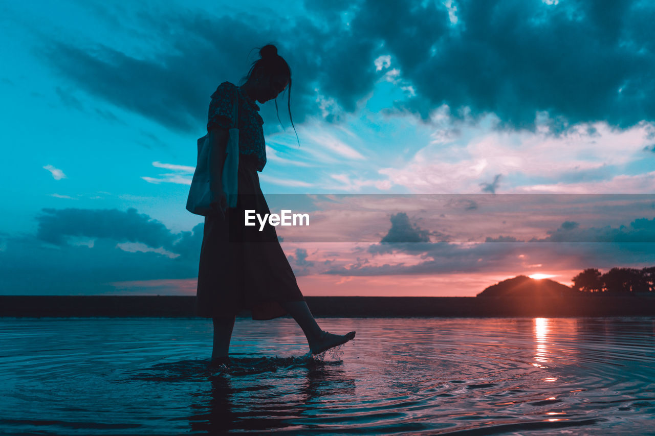 SILHOUETTE WOMAN STANDING AT SEA SHORE AGAINST SKY DURING SUNSET