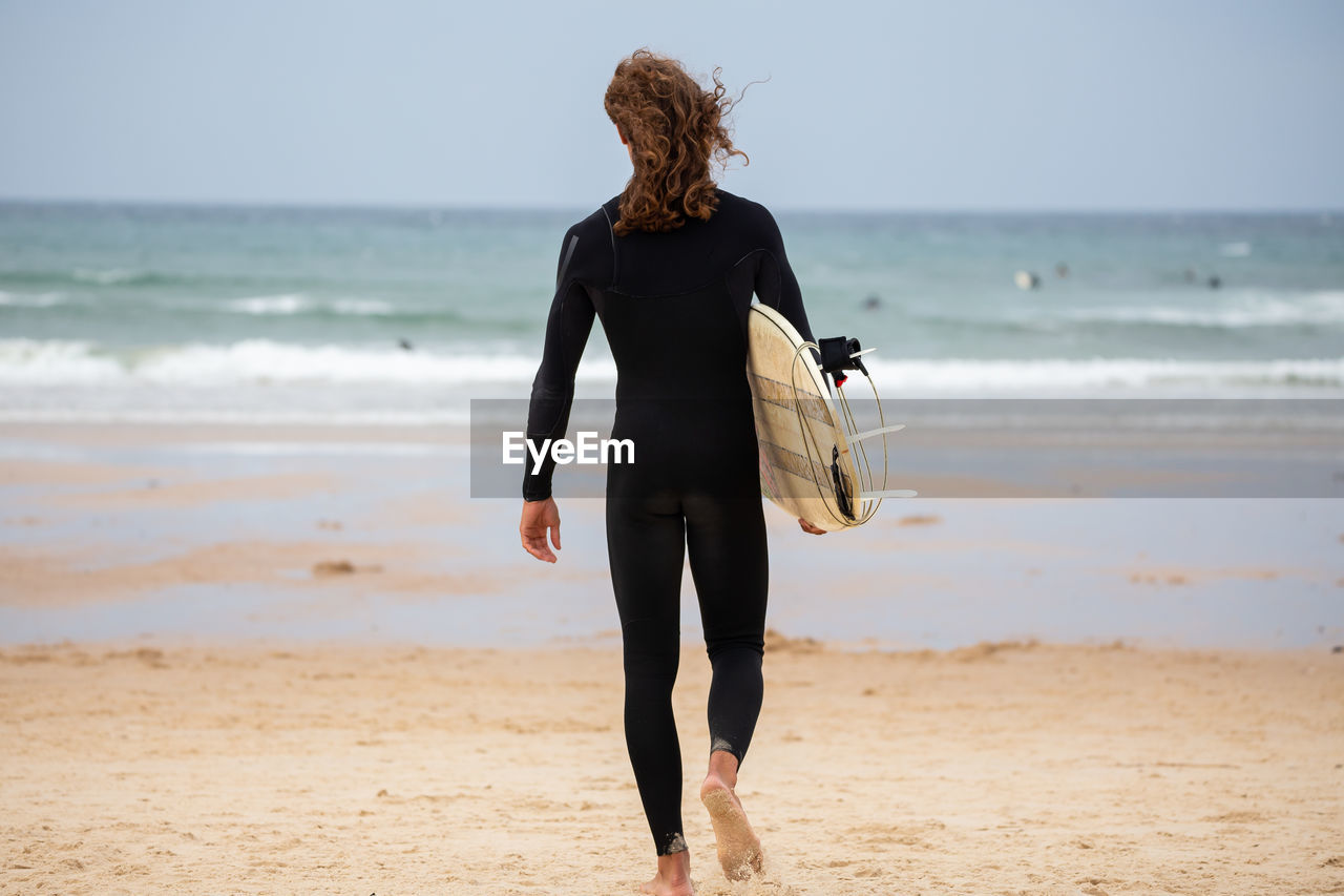 REAR VIEW OF WOMAN WALKING ON BEACH AGAINST SEA