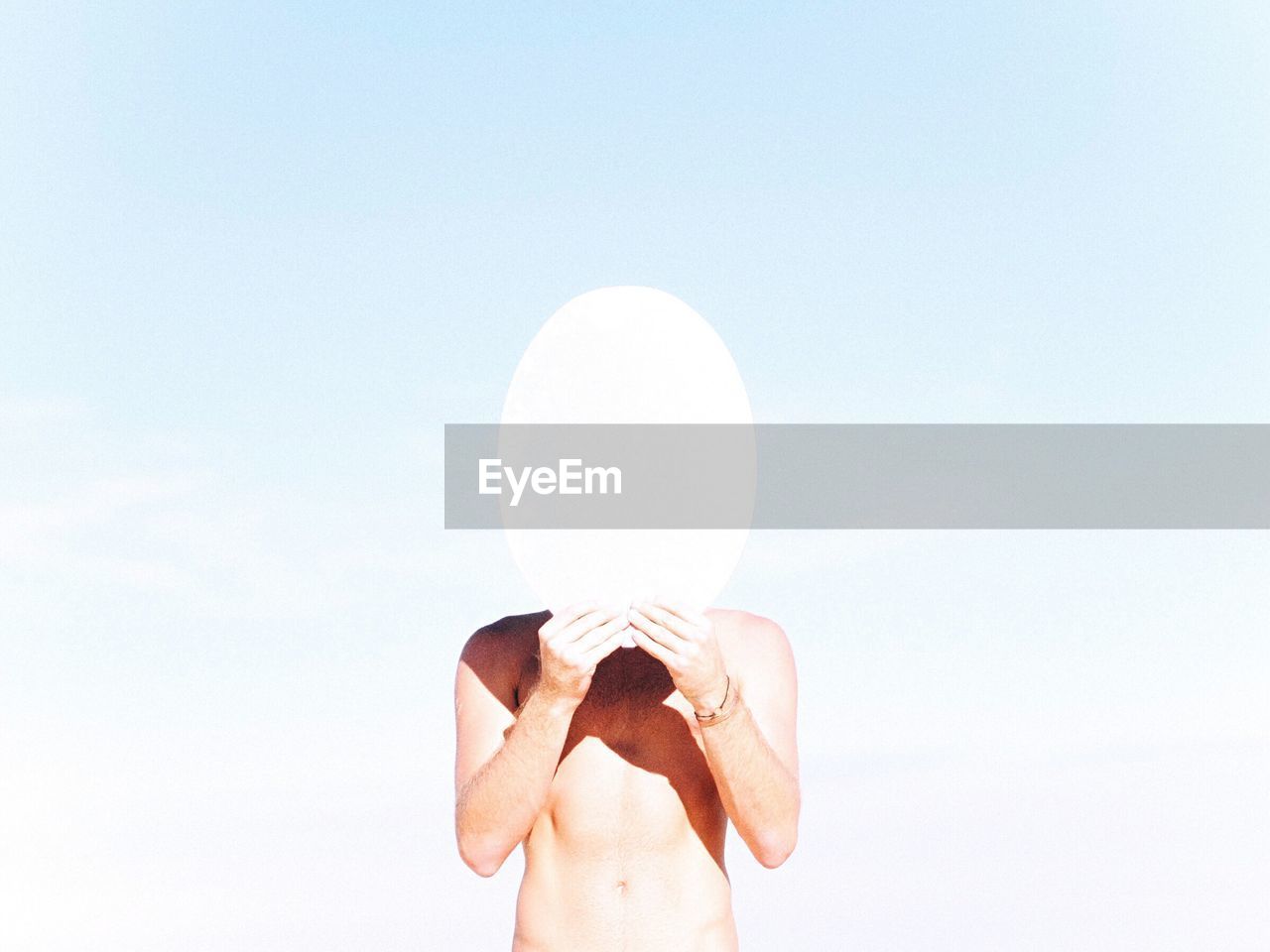 Shirtless man covering face with ellipse against sky