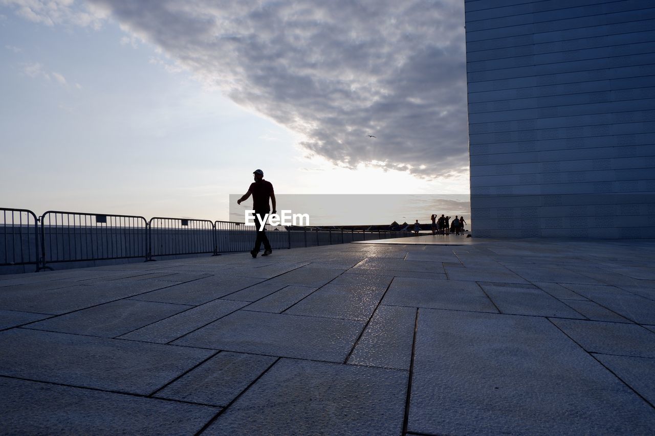 sky, horizon, architecture, one person, full length, blue, morning, silhouette, adult, city, built structure, nature, men, cloud, lifestyles, reflection, walking, sunlight, footpath, building exterior, standing, walkway, outdoors, city life, leisure activity, day, light, line, sea, rear view, shadow, road surface