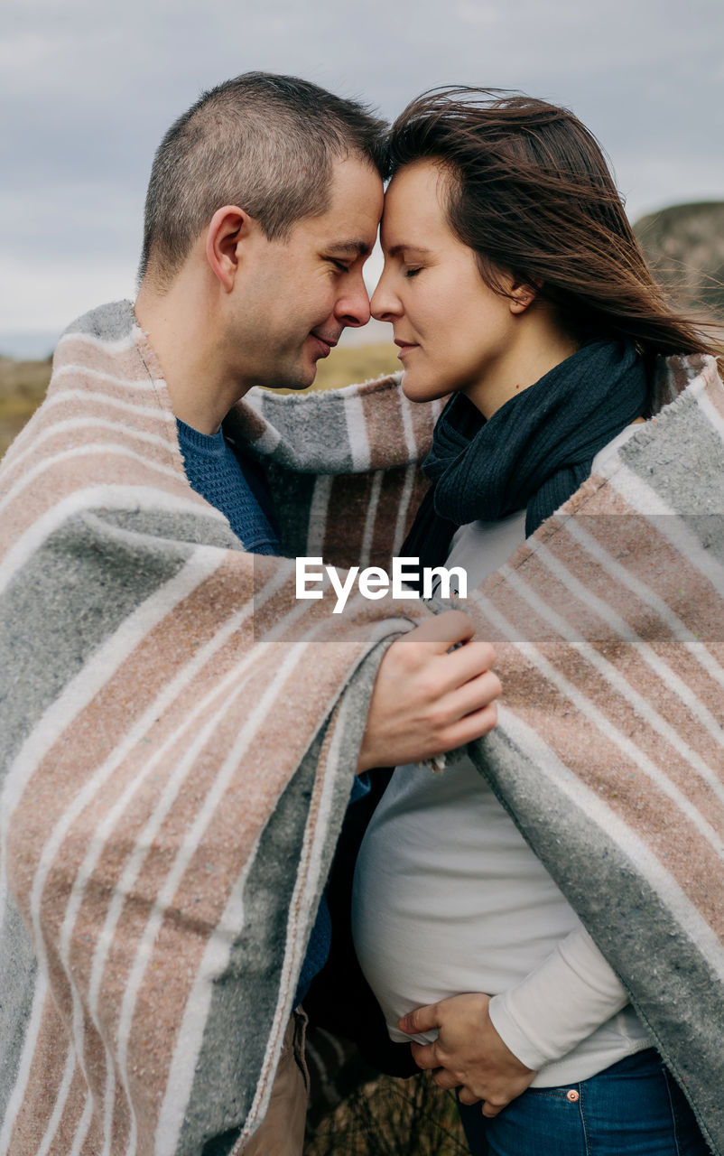 Pregnant woman with man wrapped in blanket while standing on field against sky