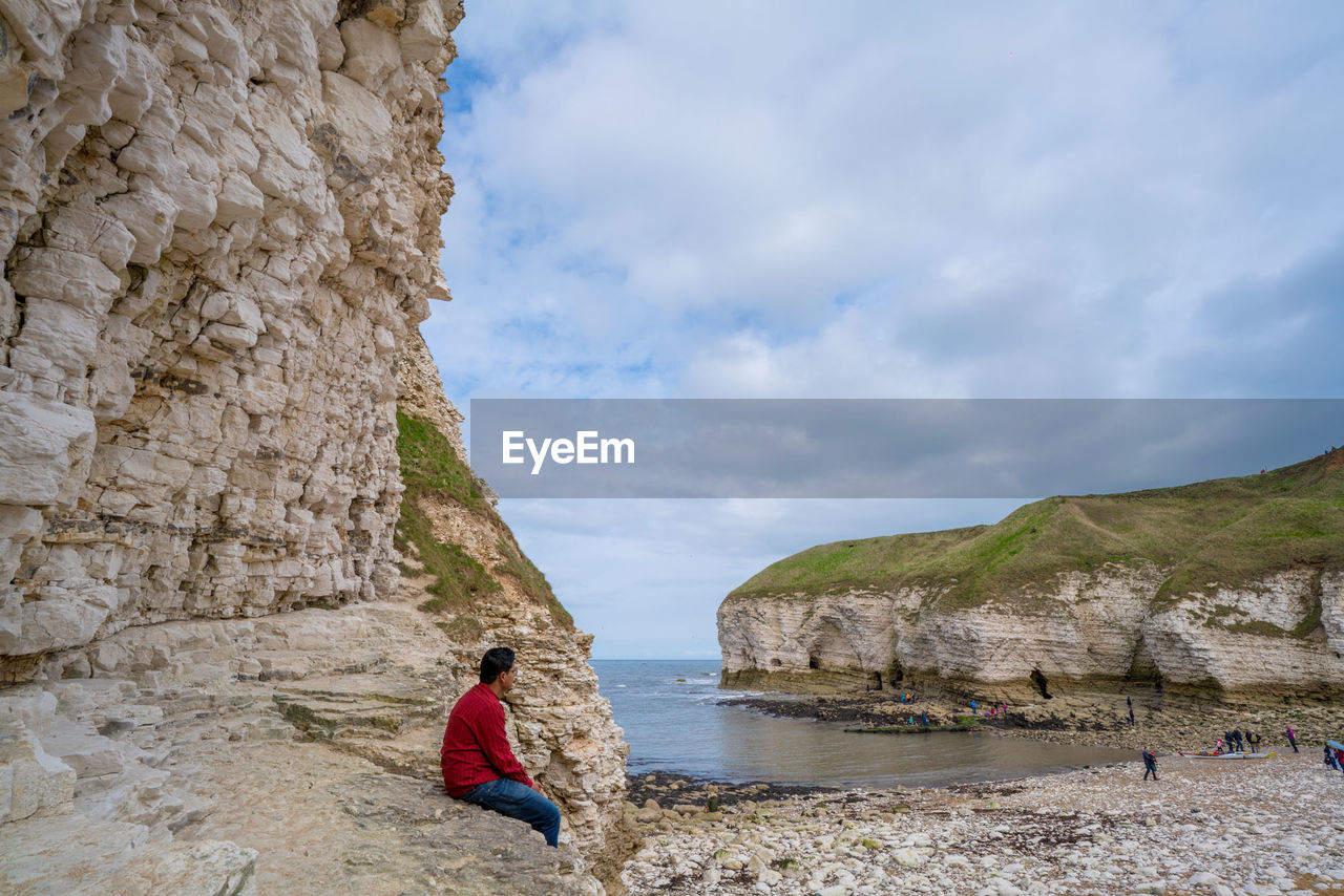 Side view of man sitting on rock at beach against cloudy sky