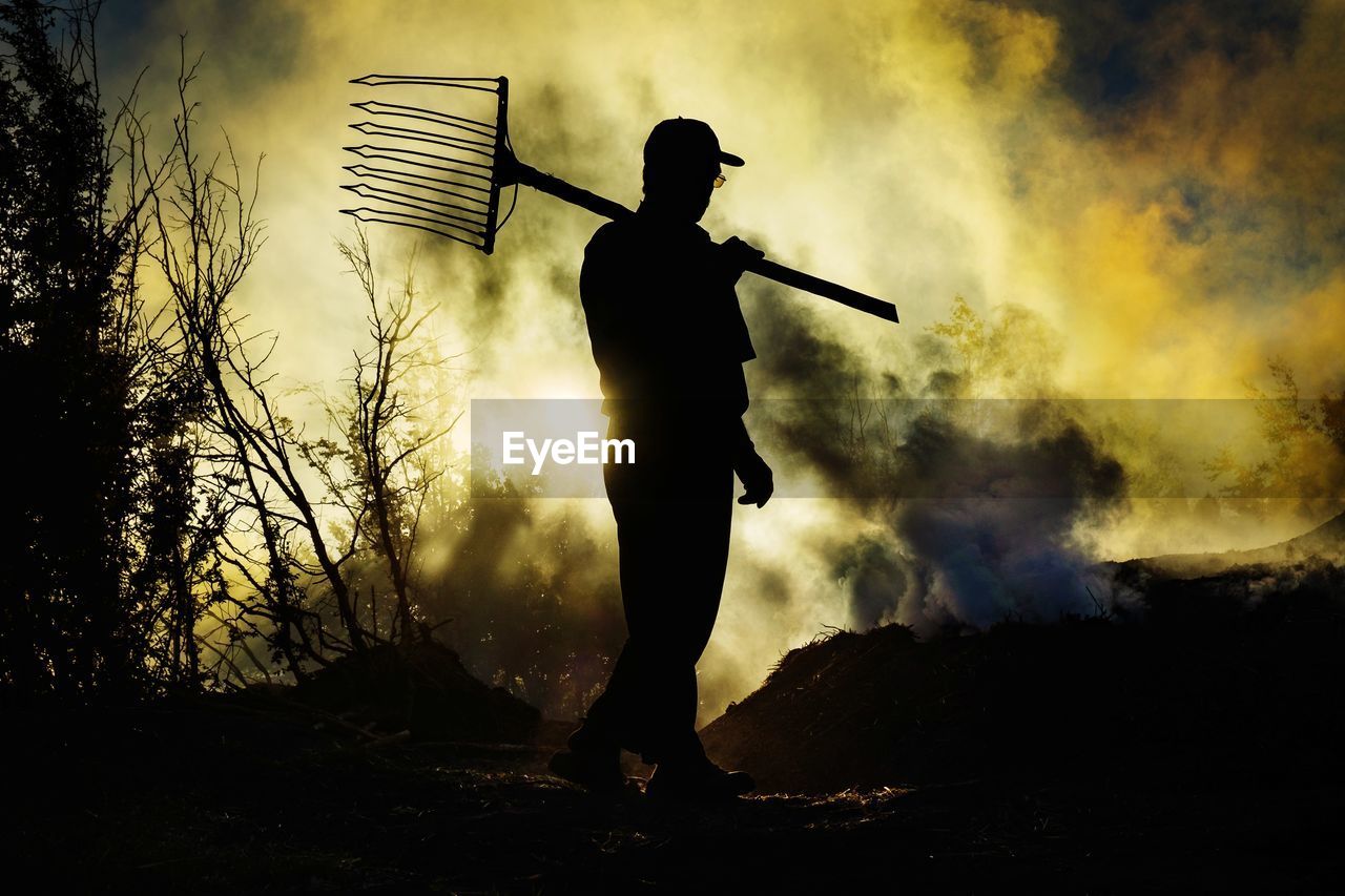 Silhouette man walking with pitchfork against sky during sunset