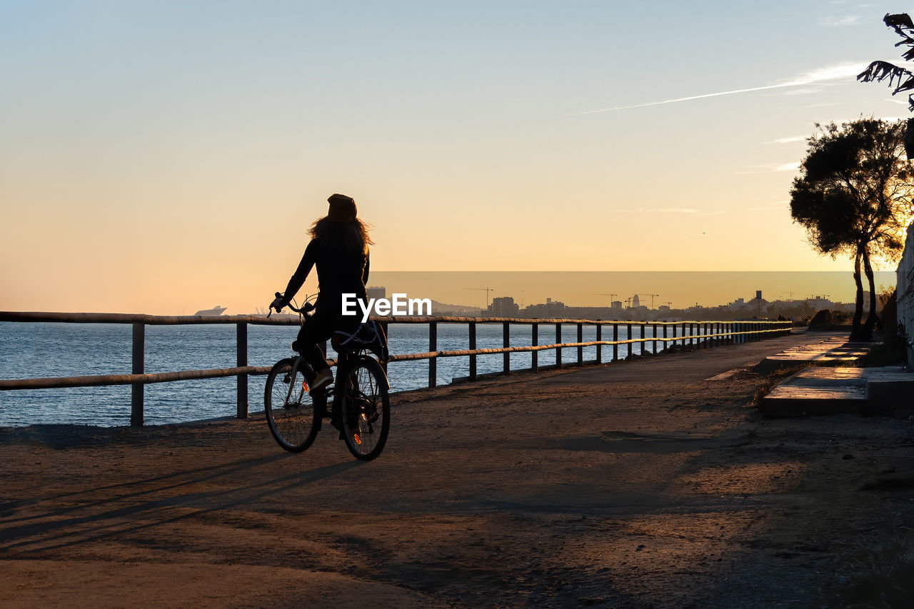 Silhouette woman riding bicycle on promenade against sky during sunset