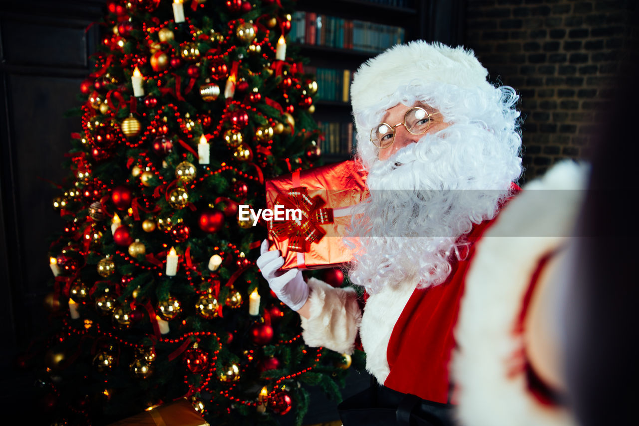 Portrait of santa clause holding gift by christmas tree at home