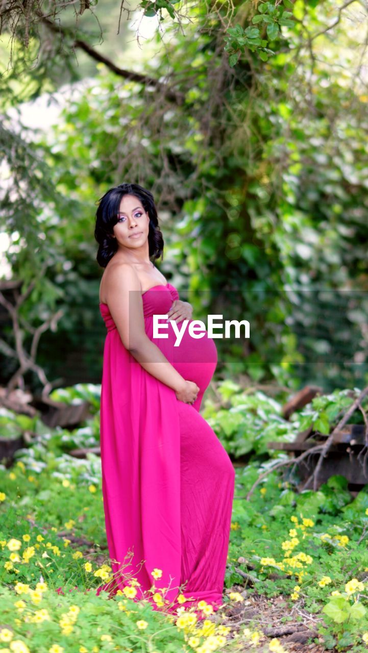 Portrait of pregnant woman with hands on stomach standing against trees
