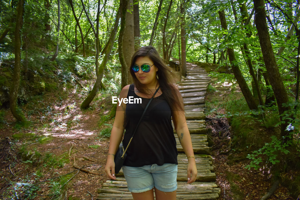 Mid adult woman wearing sunglasses while standing on boardwalk amidst trees in forest