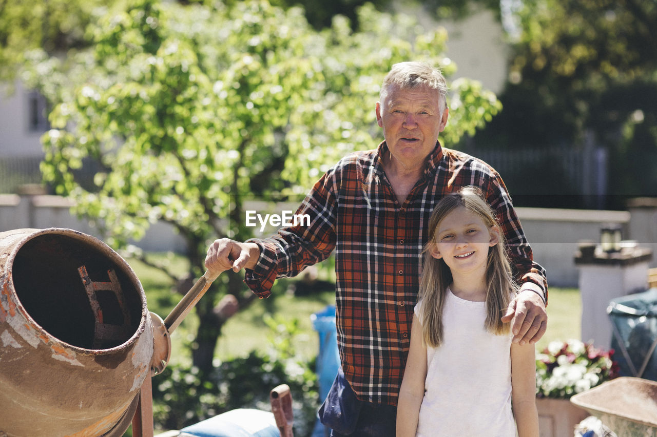 Portrait of confident grandfather and granddaughter standing by cement mixer in yard