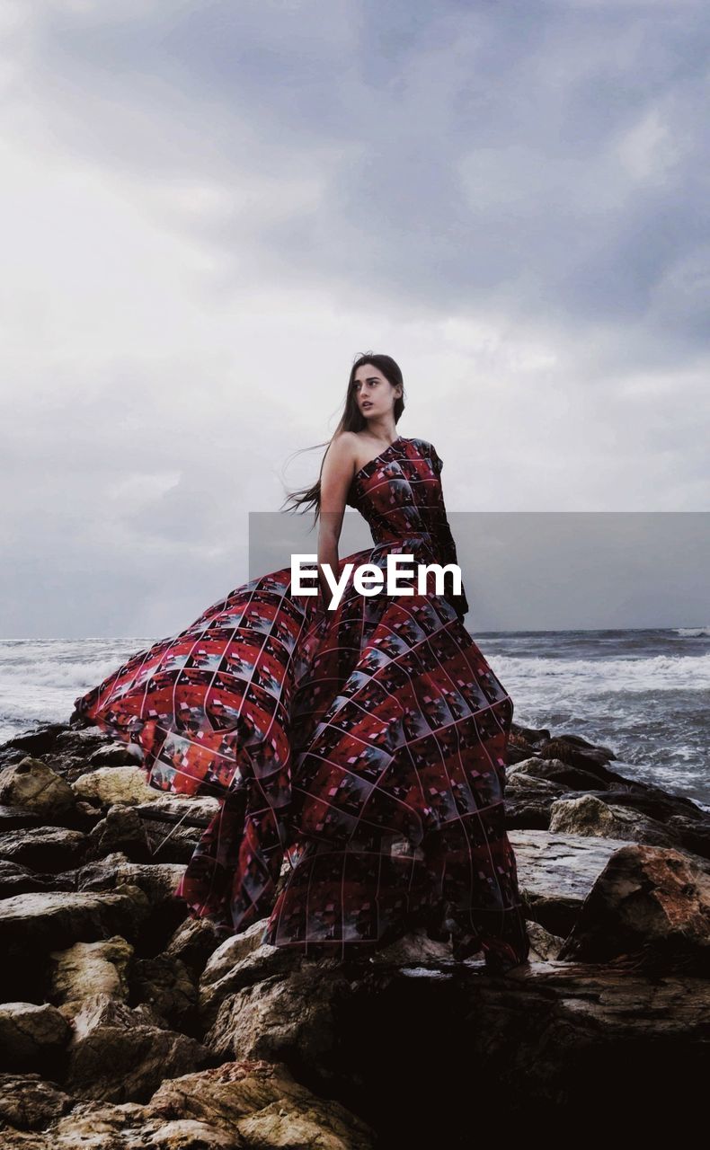 Portrait of young woman on rock at beach against sky