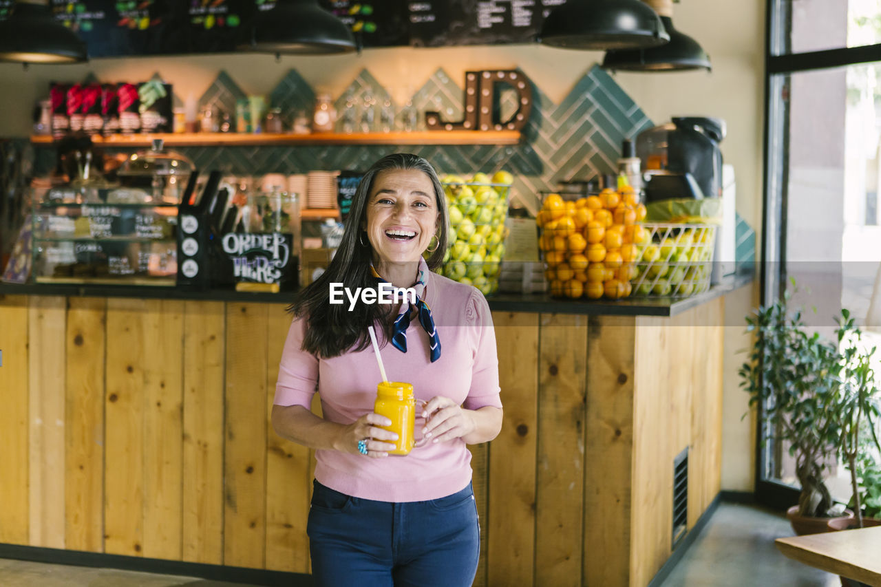 Cheerful mature woman holding mason jar with fresh juice while walking in cafe