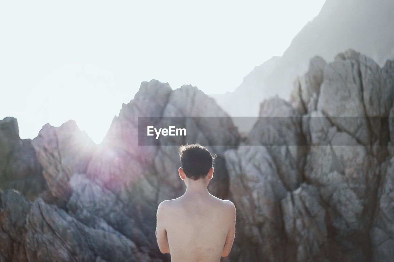 Rear view of shirtless man standing by rocks against sky