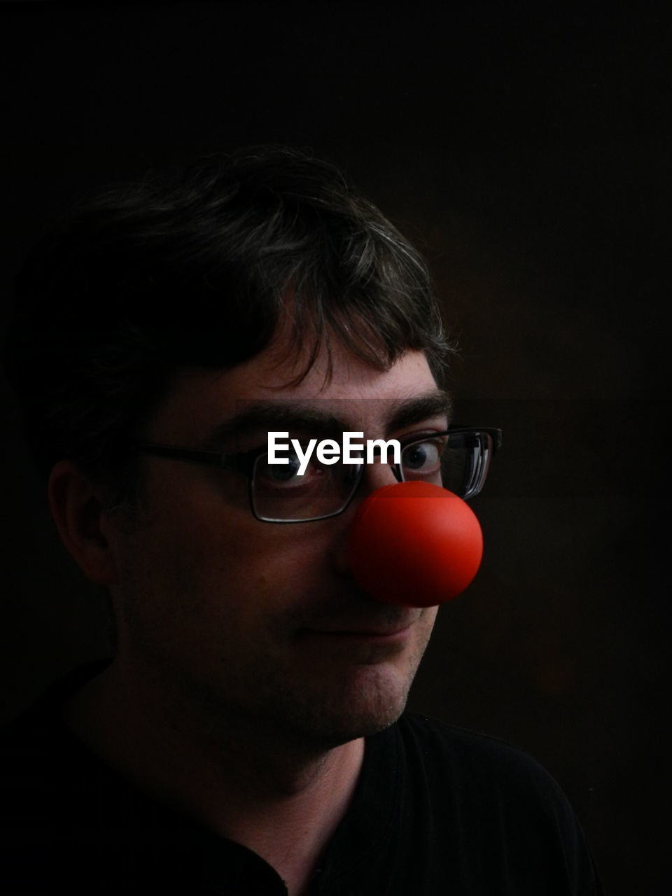 Portrait of man wearing eyeglasses with clown nose against black background