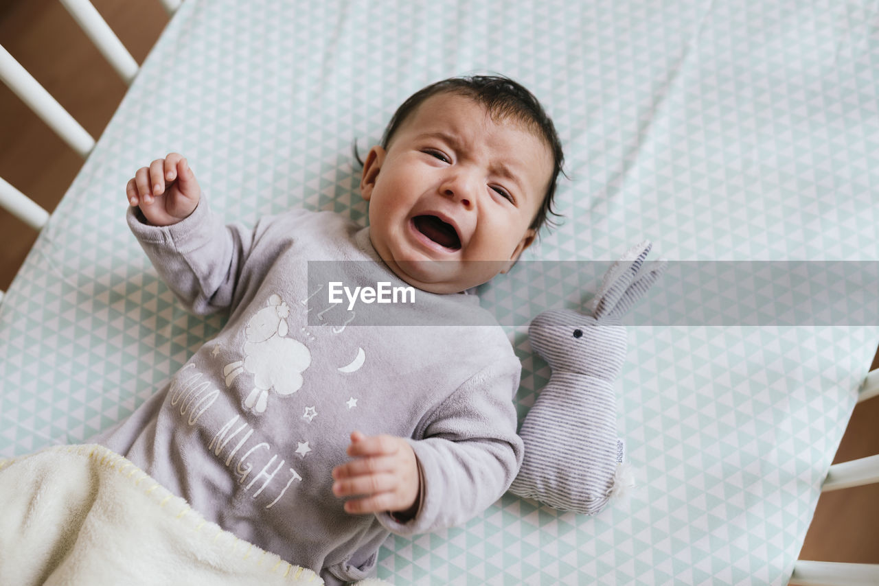 Portrait of crying baby girl lying in crib with toy bunny