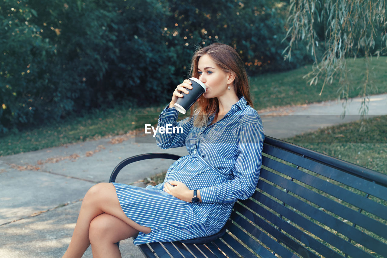 Pregnant woman drinking coffee while sitting on bench at park