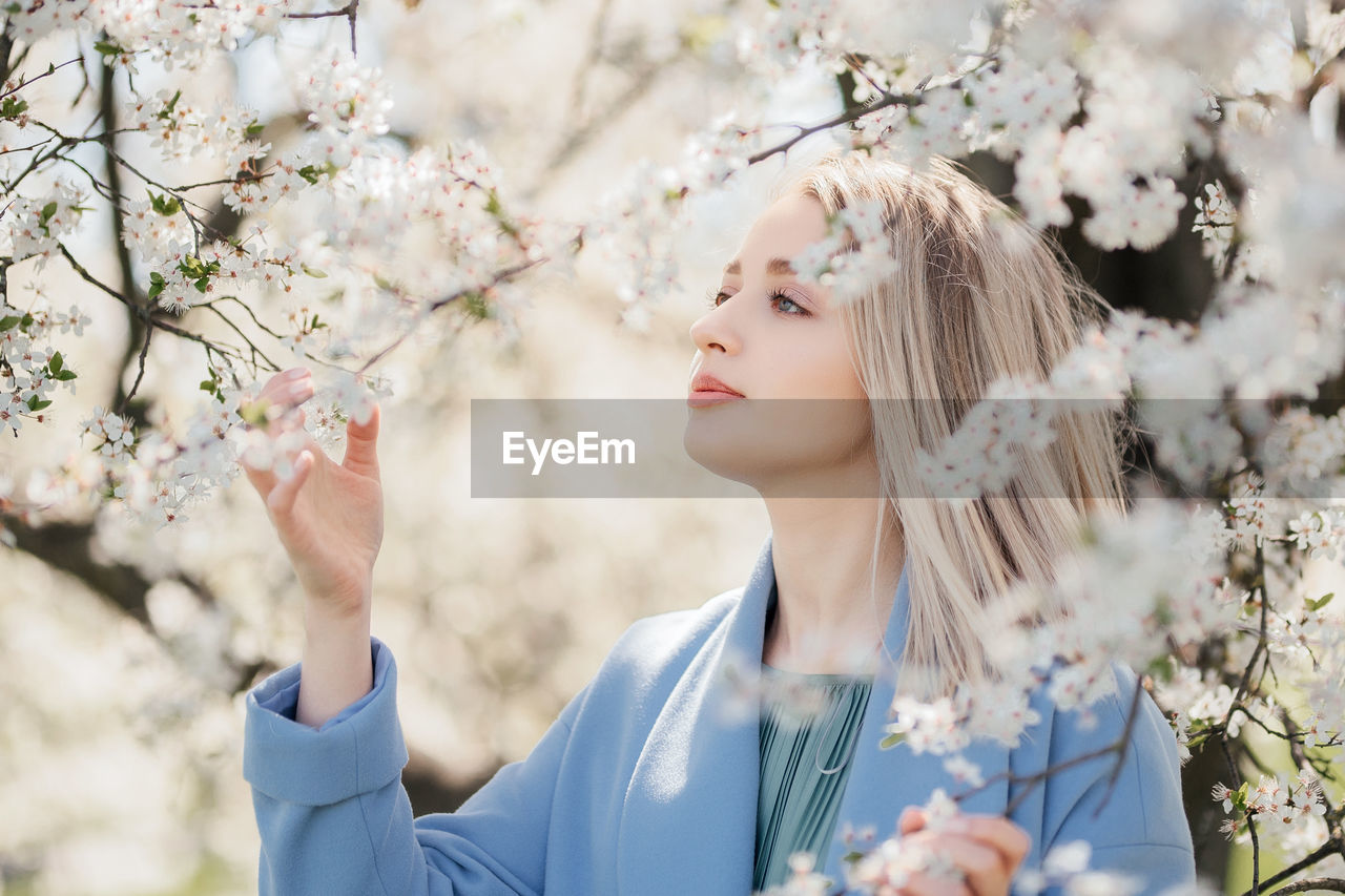 Portrait of young beautiful blonde woman posing in blooming white apple flowers on branches