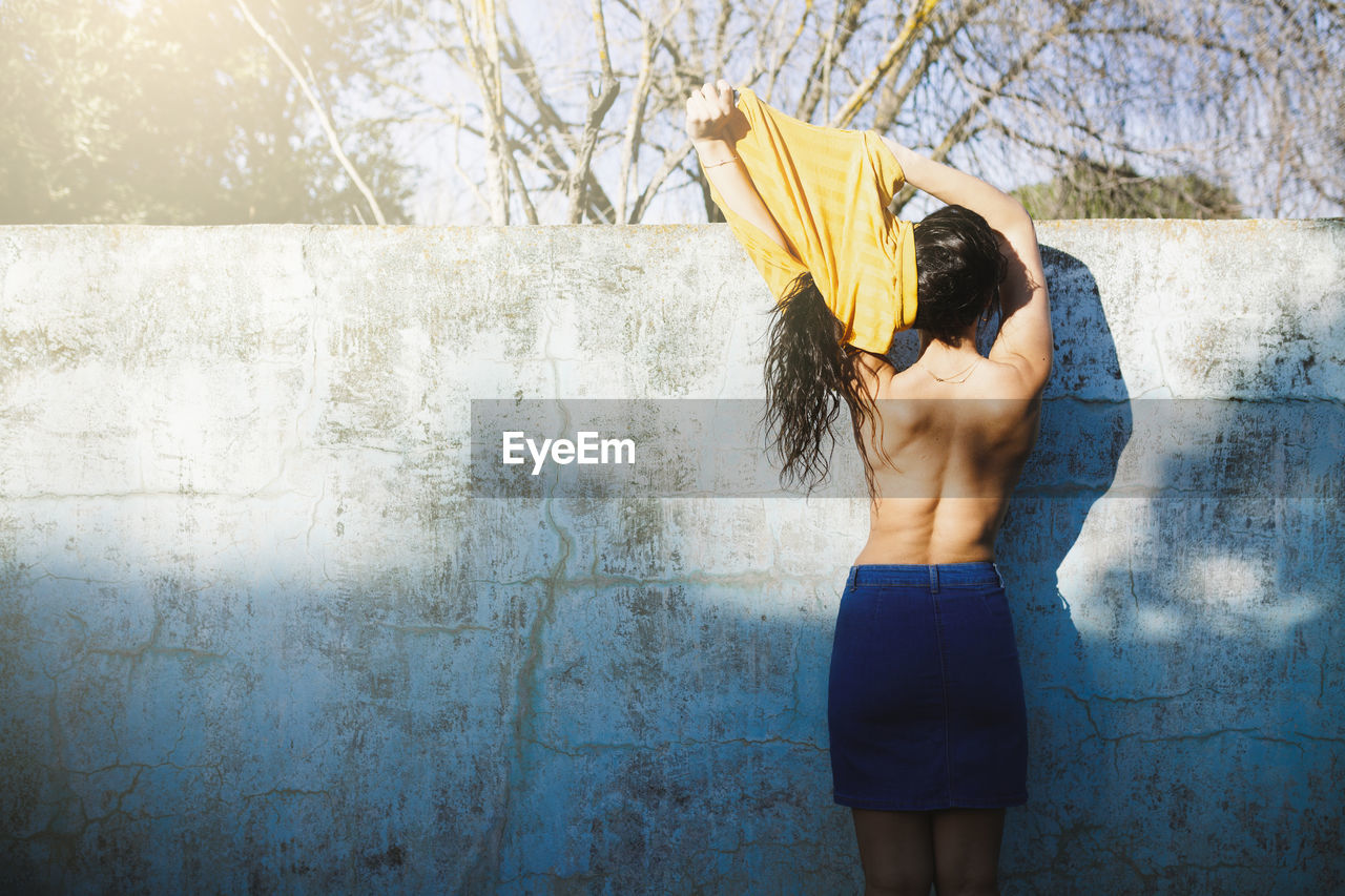 Rear view of shirtless woman removing top while standing against wall