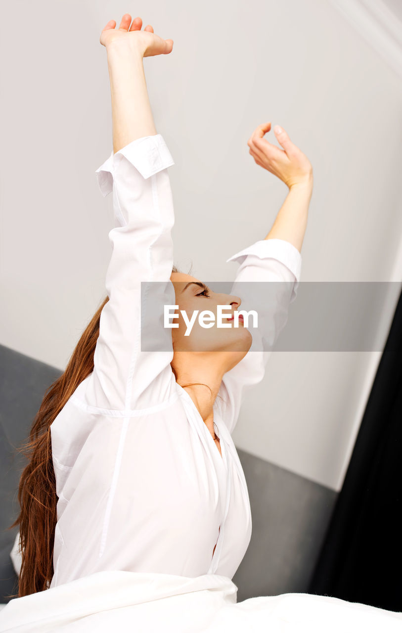 Mid adult woman in white shirt stretching arms on bed