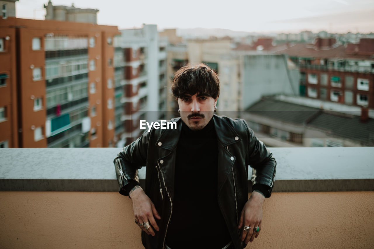 Serious man in leather jacket standing on rooftop and leaning on fencing and confidently looking at camera