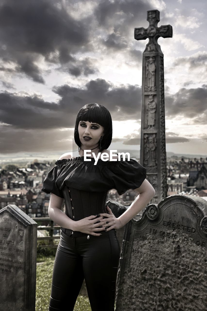 Portrait of goth woman standing in cemetery against sky