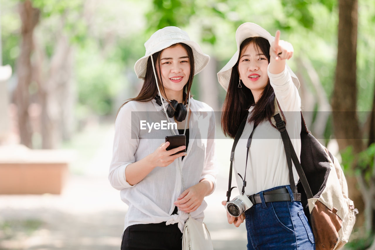 Portrait of smiling young woman using mobile phone by friend pointing against trees