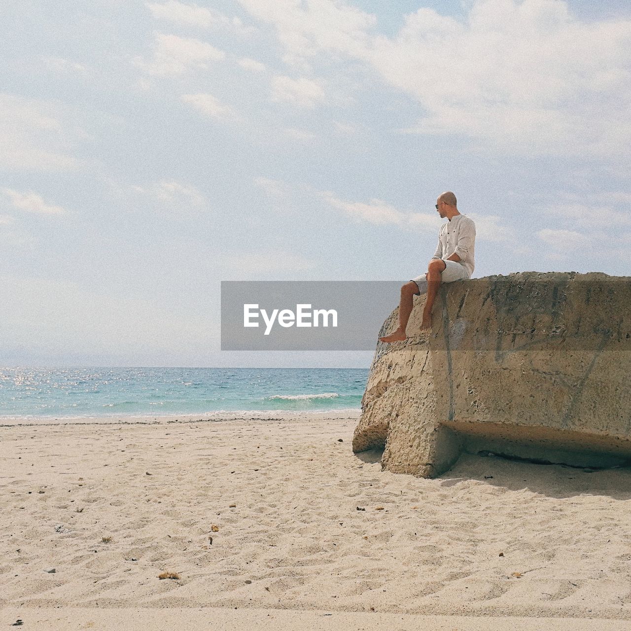 Low angle view of man sitting on rock at beach against cloudy sky