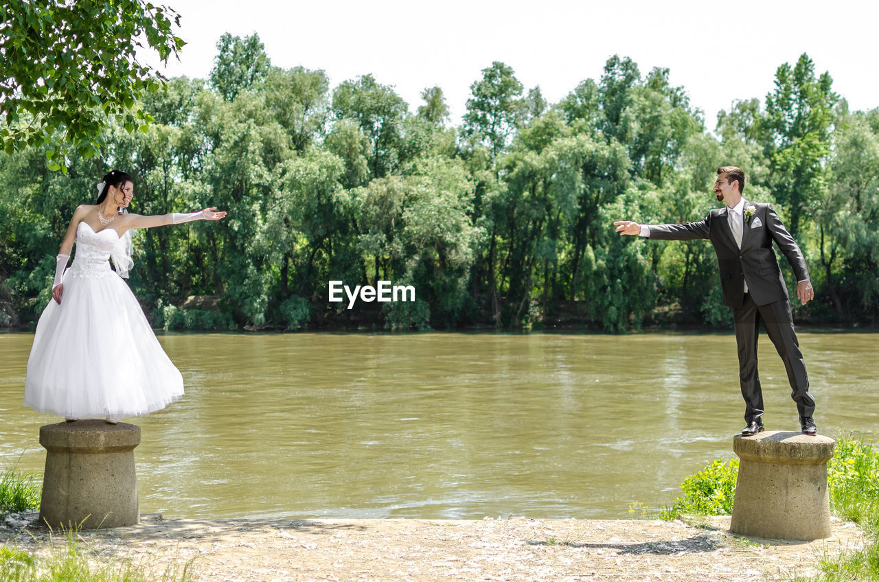 Full length of wedding couple gesturing while standing on bollard against river