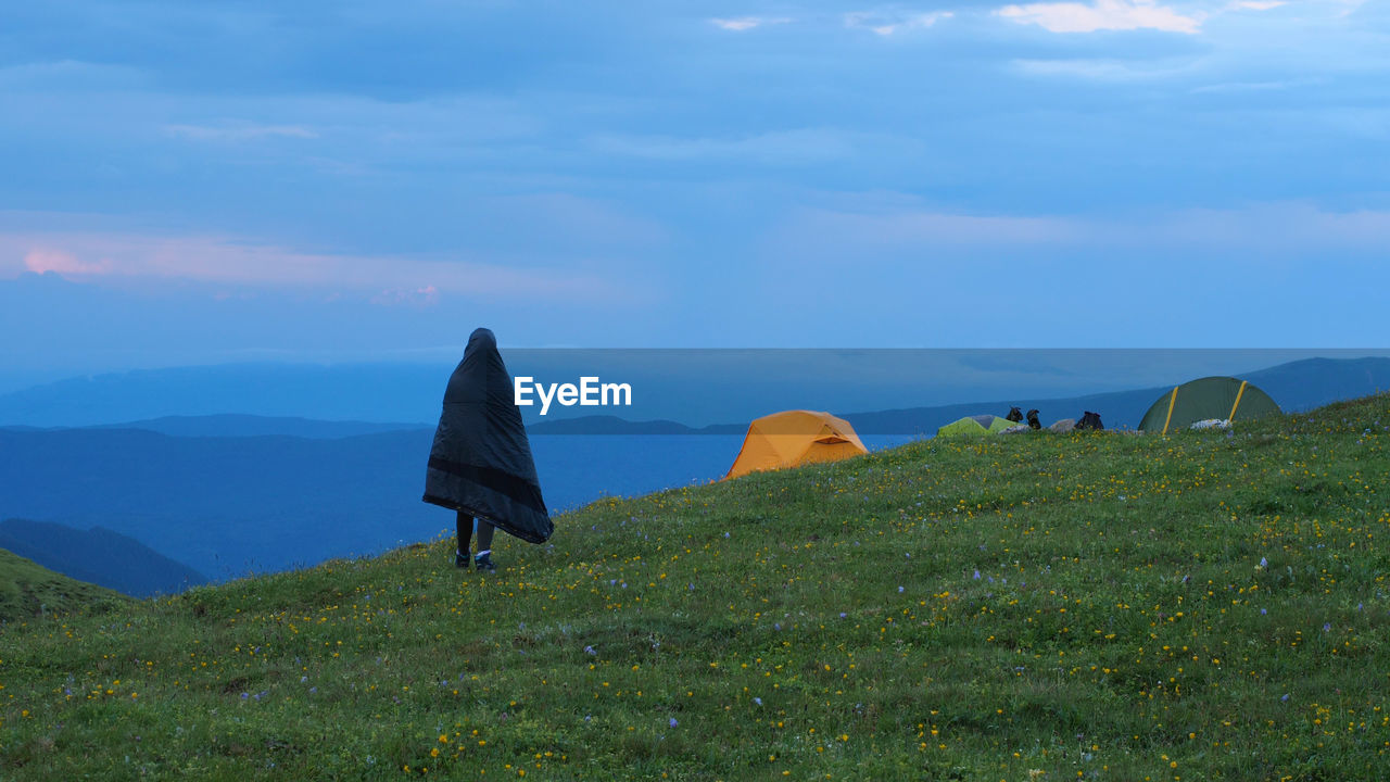 A hiker wrapped up in a sleeping bag walking across green grass on a background of mountain peaks