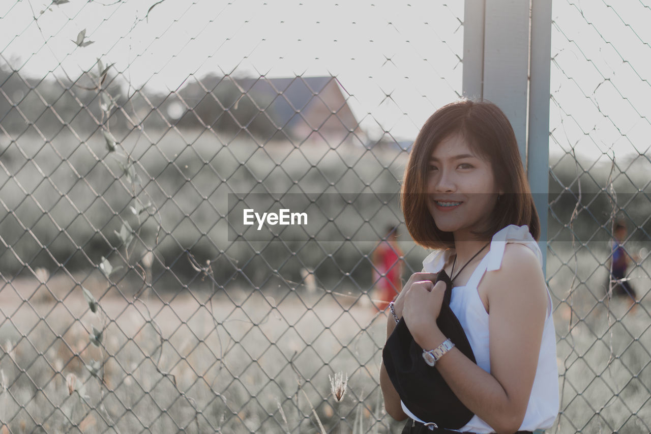 Portrait of young woman standing against chainlink fence