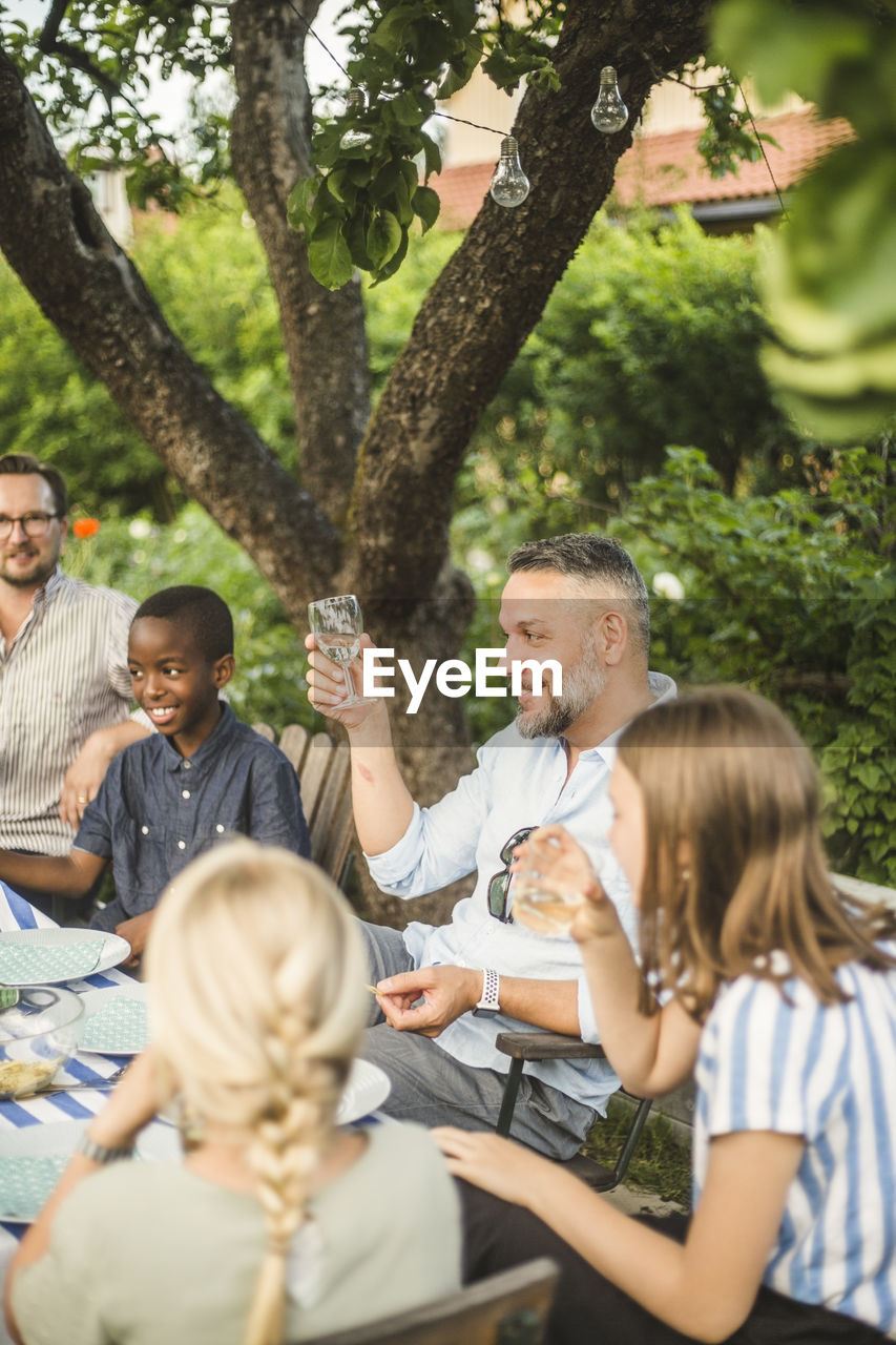 Mature man raising wine glass while sitting with friend and children in backyard party